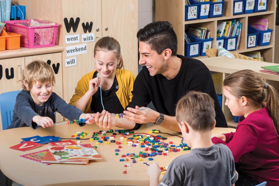 A student teacher sits at a table with four young children smiling and making crafts.