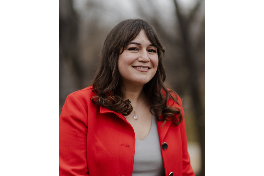 head shot of woman with dark long hair with bangs, wearing red coat, trees in background