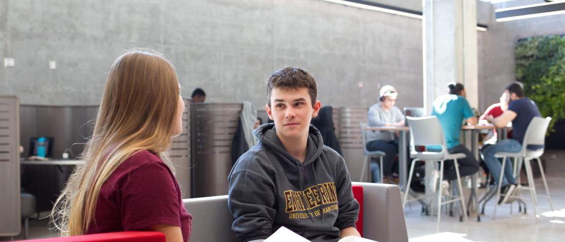 Two students seated together studying, several other students are also in the background studying.