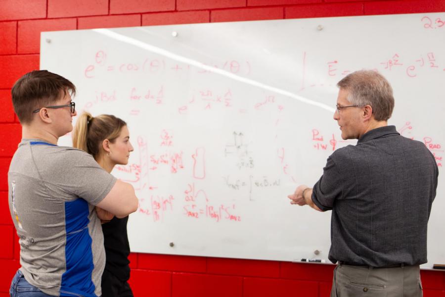 Two students stand at a white board while a professor speaks.