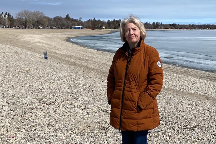 Doctor mary benbow in brown jacket standing on a beach with a lake behind her.