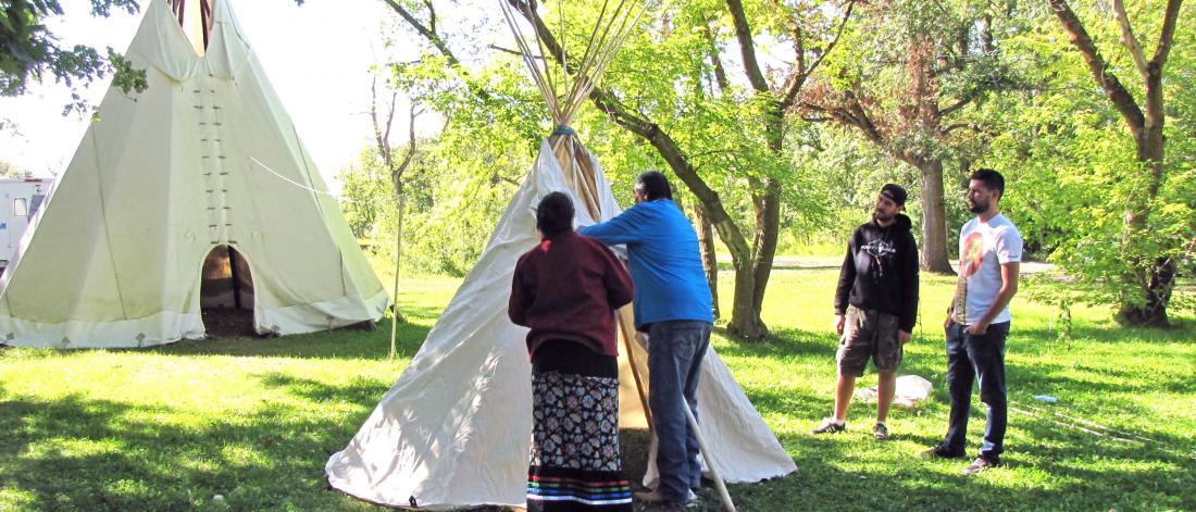 Two Indigenous elders work on making a teepee tent while two students observe.