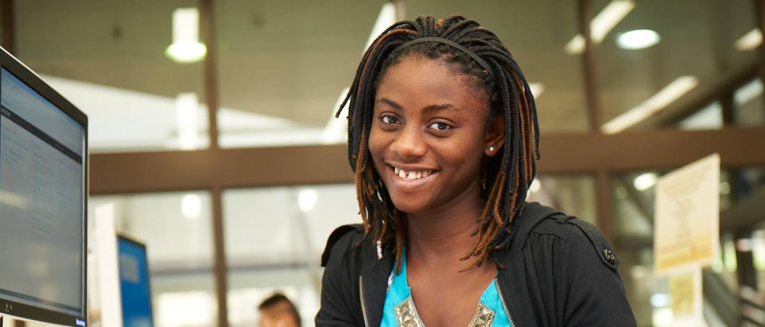 A students smiles as she sits at a computer work station in a classroom.