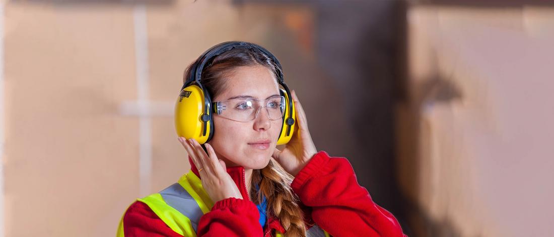 Woman in safety vest wearing yellow safety headphones with hands placed on the headphones.