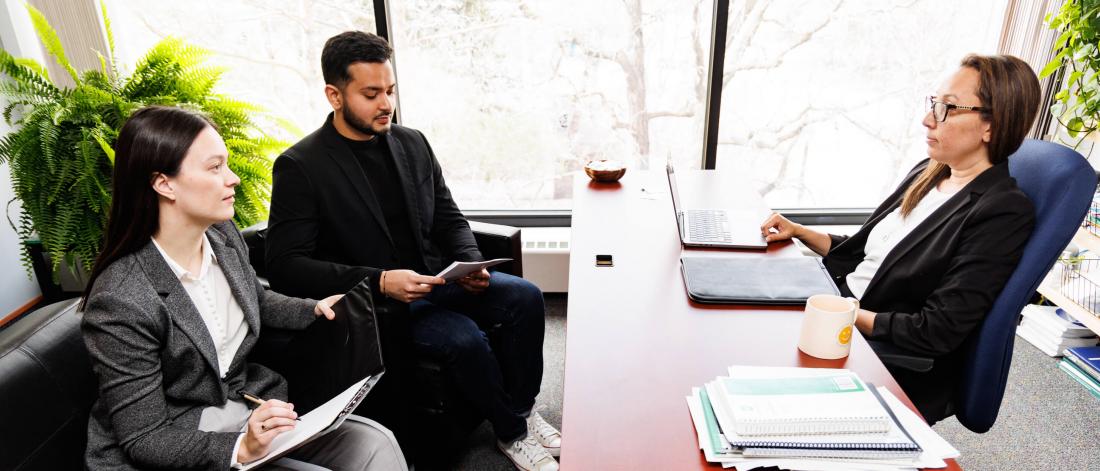 Two students in suits meet across a desk with a professor also wearing a suit