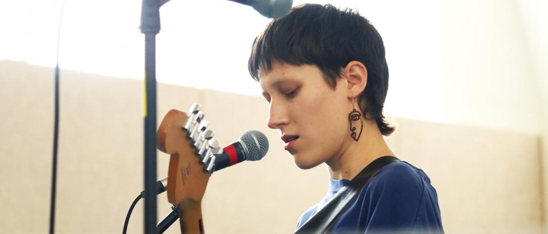 Person with short hair holding a guitar speaks into mic