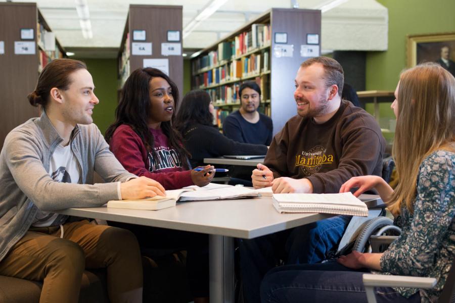 A diverse group of students sit at a table together in a library studying.