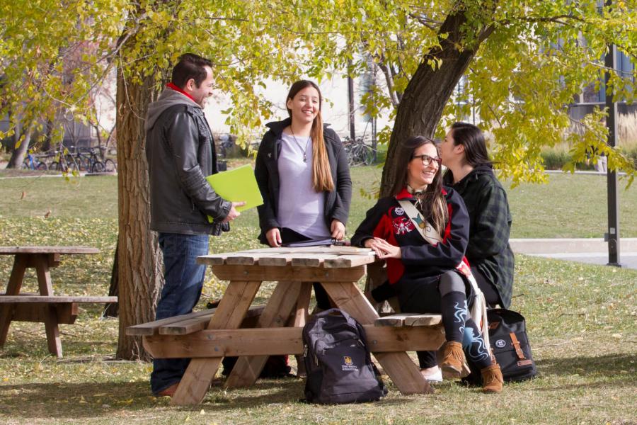 Four Indigenous students sit and stand together talking outdoors at a picnic table. 
