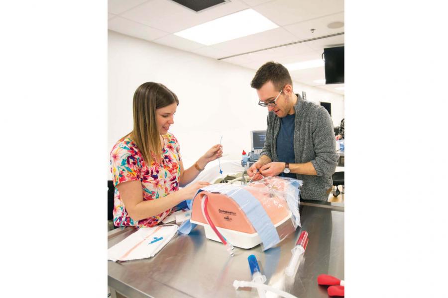 Two medical students practice administering a medical drip to a practice mannequin.