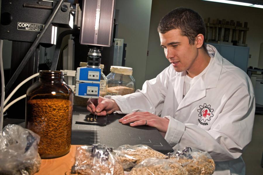 A graduate student wears a white lab coat and sits at a table working in a lab. 