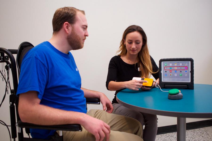 An occupational therapist sits at a small table with a patient and points to a large yellow button in front of a tablet.