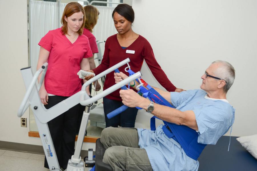 Two occupational therapists use a machine operated lift to help a patient out of bed.