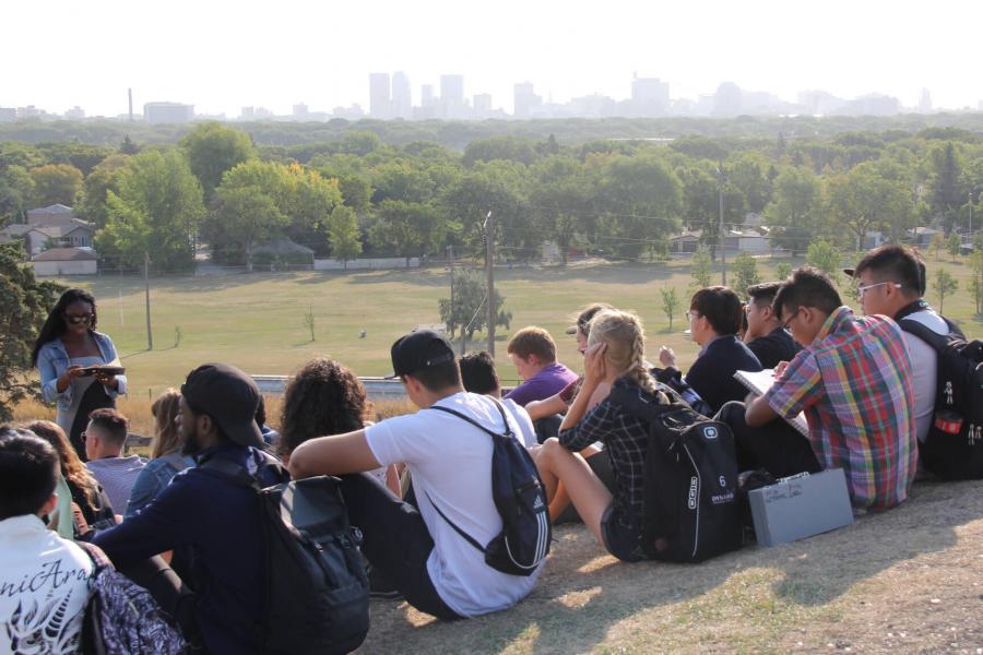 Students seated on the ground at the top of a hill overlooking a scenic city landscape.
