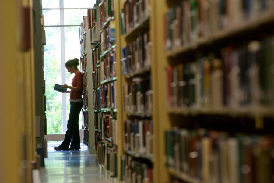 A student leans against a shelf of books while reading. 