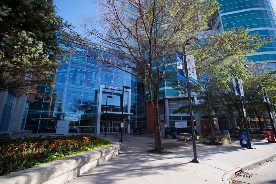 A wideshot of the Bannatyne campus in the fall. The sky is blue and the trees are mostly green, but they've shed a few leaves. A flowerbed with red flowers decorates the entrance to the building.