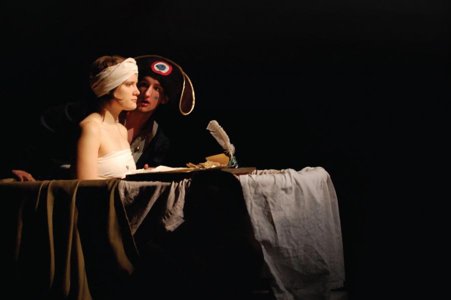 Two student performers on stage with black background. One student is looking off into the distance while sitting in a bathtub.