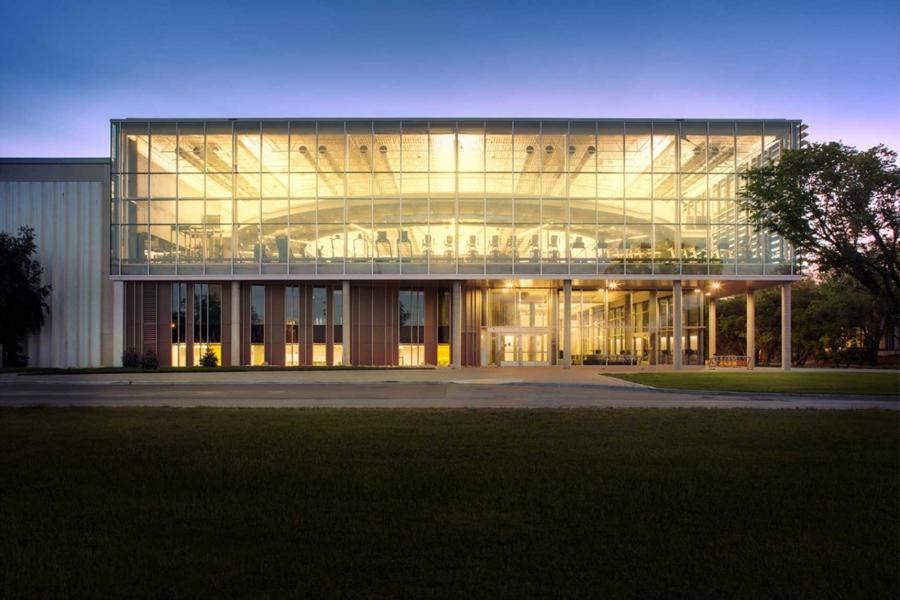 Exterior of the Active Living Centre at night. Large glass walls reveal a warmly lit interior with a walking track, several treadmills and other exercise equipment.
