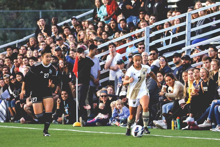 Two women in jerseys intensely play soccer on an outdoor pitch while bleachers full of cheering spectators look on.