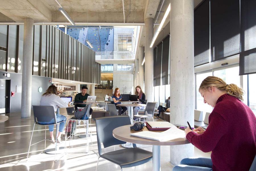 Five students with notebooks and laptops sit working at small round tables in a brightly lit room with high ceilings and concrete floors and pillars. A climbing wall within the Active Living Centre is in the background.