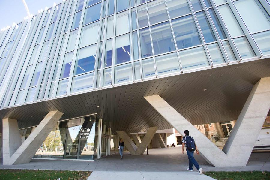 Two students walk toward the entrance of the art lab building. The building is a modern design with a glass and metal exterior appearing to be suspended on diagonal concrete pillars.