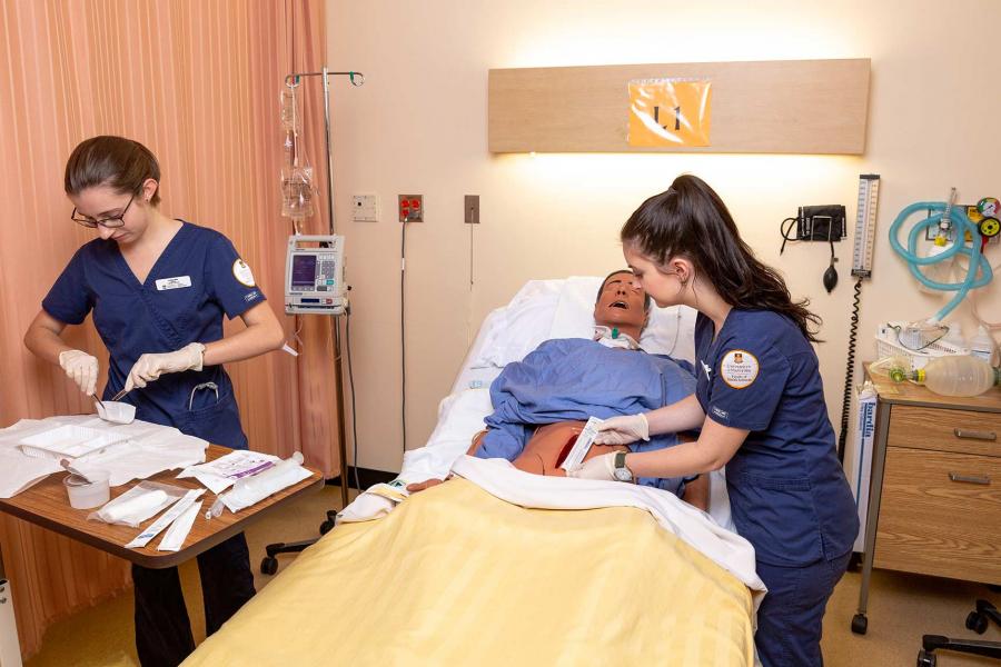 Two students wearing scrubs in a hospital room perform a procedure on a mannequin in a hospital bed with its abdomen exposed.