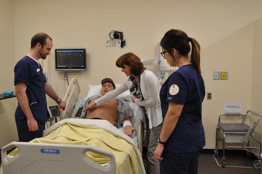 Two students in scrubs and an instructor in a hospital room standing over a mannequin wearing a baseball cap in a hospital bed.