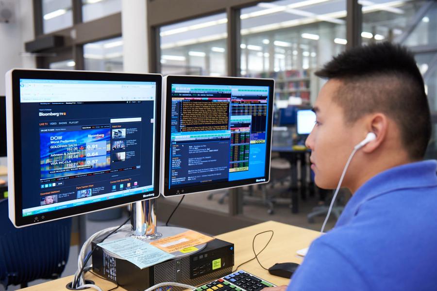 A student wearing wired headphones faces two Bloomberg computer screens displaying financial information and stocks from Bloomberg TV.