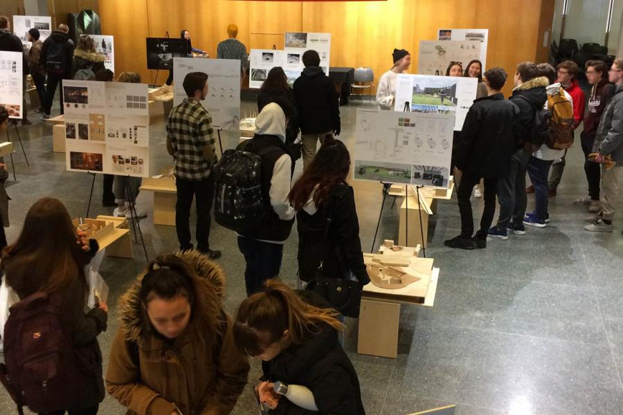 People walk around a room in the John A. Russell Building filled with architecture plans on easels and stands. The room has a sign saying ENVIRONMENTAL DESIGN in red letters on a wood panel wall in the background.