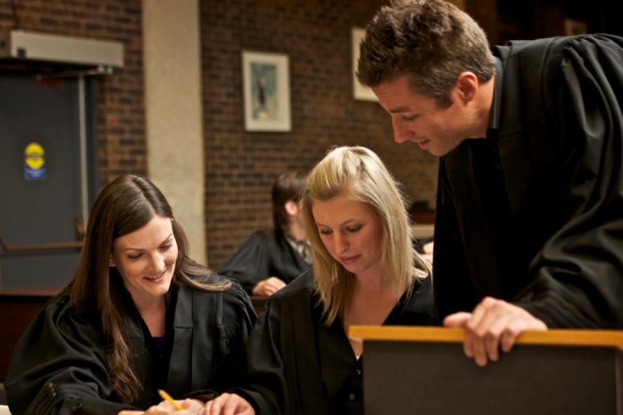 Three Faculty of Law graduates wearing black robes sign a book in a hall of the Robson Hall.