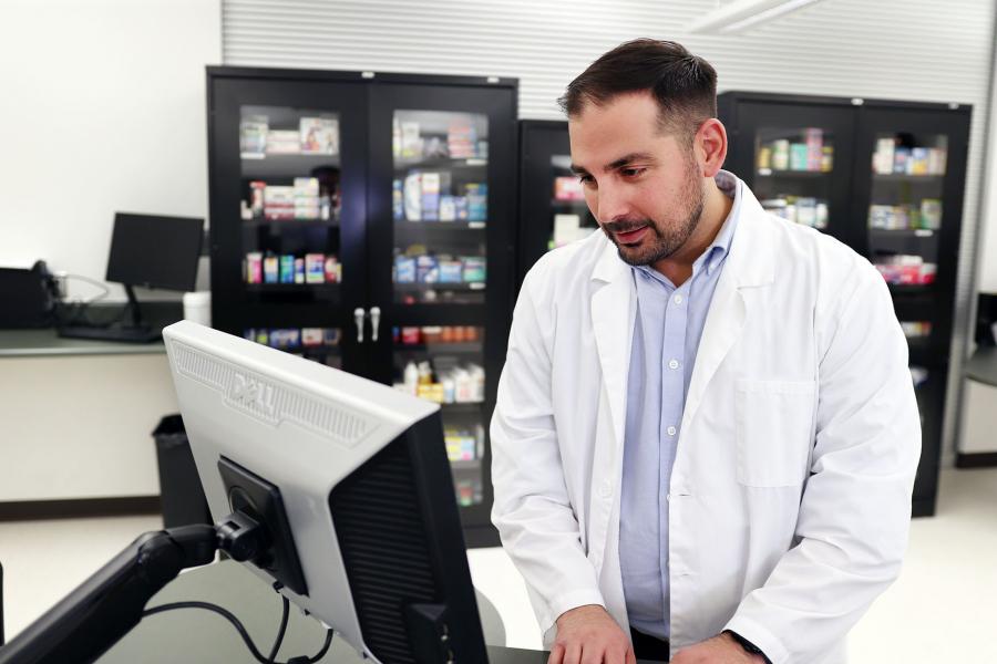 A man in a lab coat works on a computer near a cabinet with prescriptions.