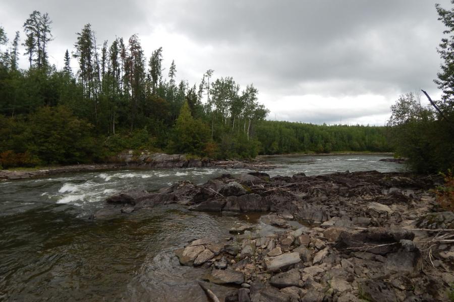 Rapids on the Laurie River, MB lined by rock and spruce forest against a cloudy sky.