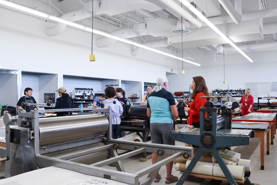 A group of people listen to a demonstration in a printmaking studio filled with equipment.