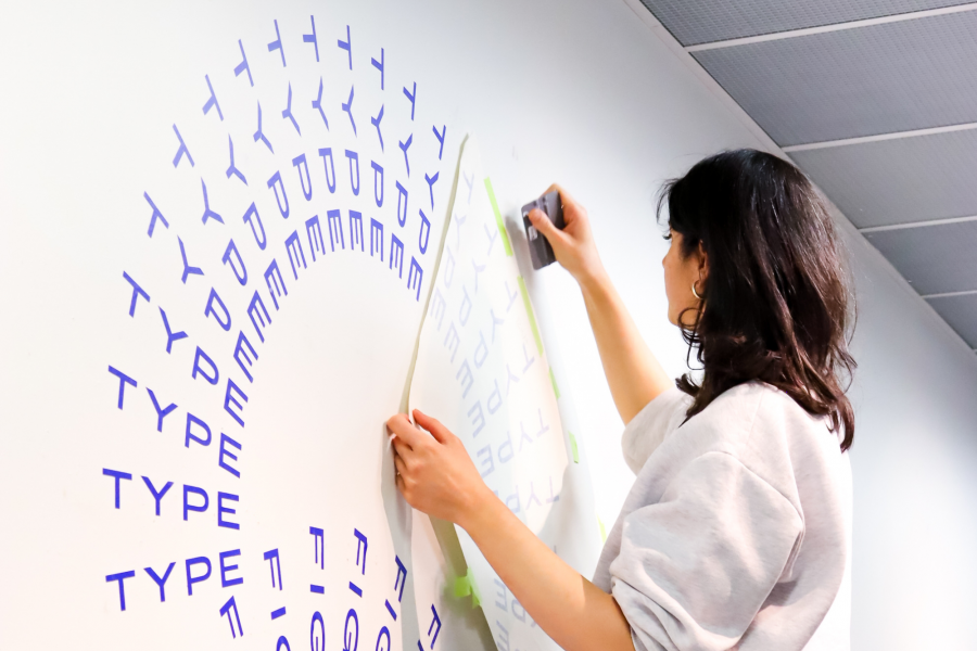 A student applies a vinyl text decal to a wall in a spiral pattern reading "TYPE".