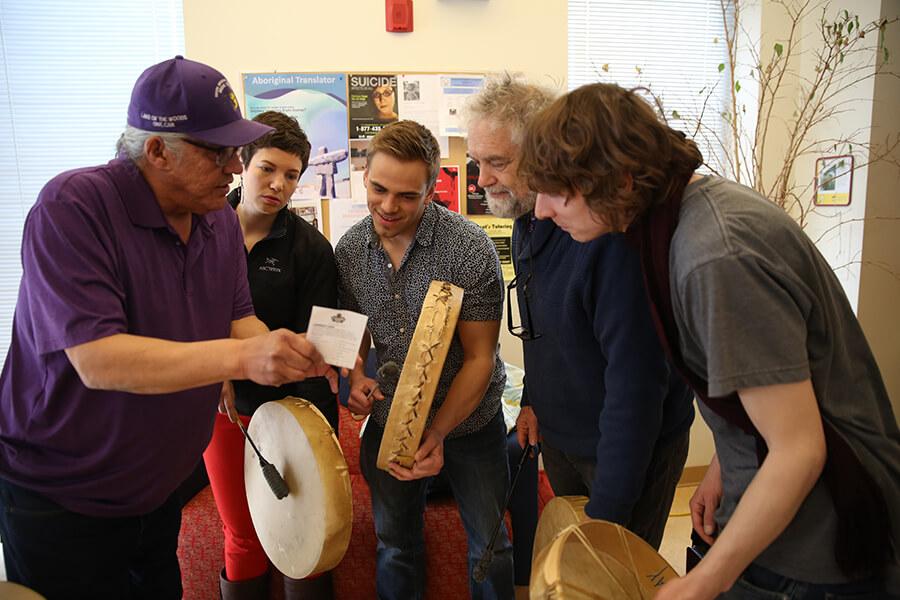 A group of five people holding Indigenous drums, huddled together looking at a card with instructions.