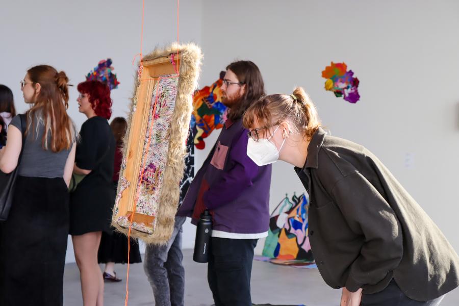 A masked viewer closely examines a suspended textile artwork during an exhibition opening.