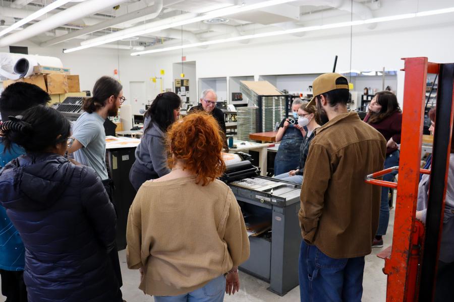 A group of students gathers around a printing press during a printmaking demonstration.