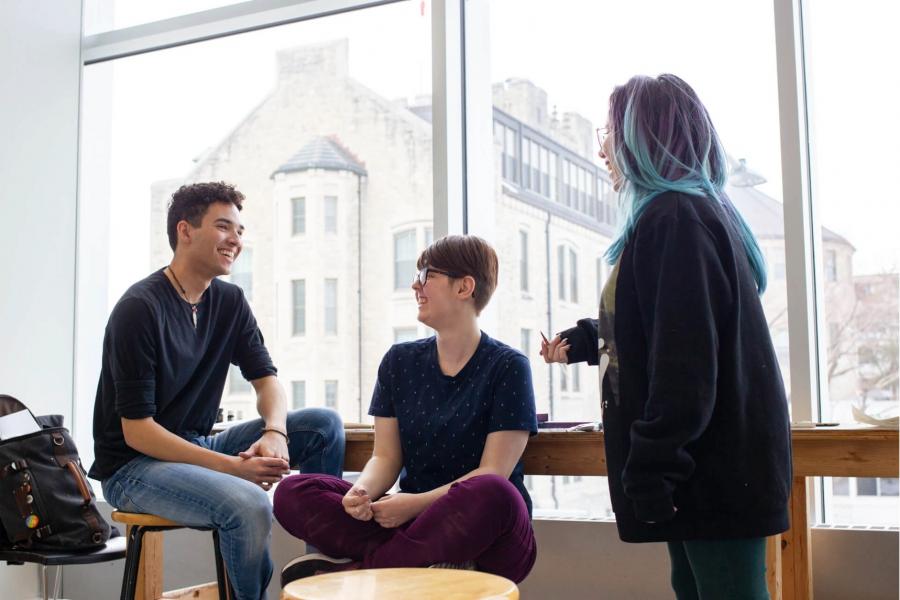 Three students sit smile together by a large window.
