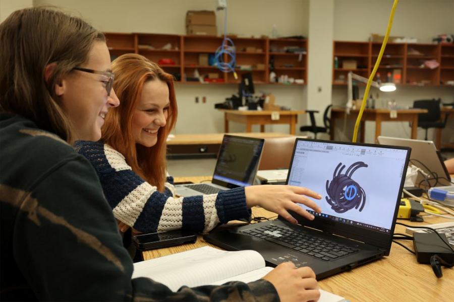 Two engineering students with laptops, one is pointing to a rendering on the other student's laptop.