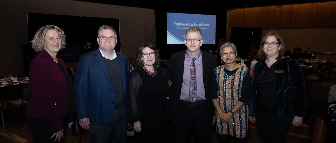 Six people stand together in a dimly lit banquet hall with round tables in the background. A presentation screen behind them displays the title ‘Empowering Excellence.