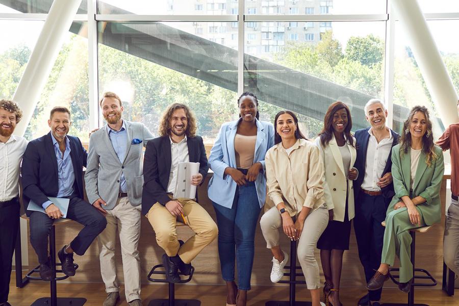 A diverse group, dressed in business casual attire, smiling and posing confidently in an office with large windows and natural light.