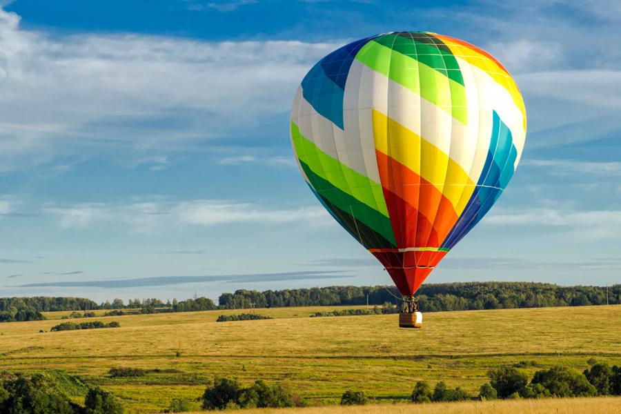 A colourful hot air balloon with rainbow stripes floats over a vast, sunlit meadow under a clear blue sky.