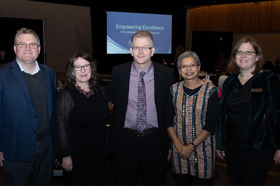 Five people stand together in a dimly lit banquet hall with round tables in the background. A presentation screen behind them displays the title ‘Empowering Excellence.