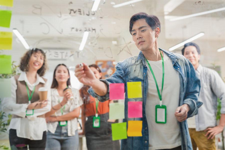 A group brainstorming session with a man writing on a transparent board covered in diagrams and notes while three colleagues watch attentively. 