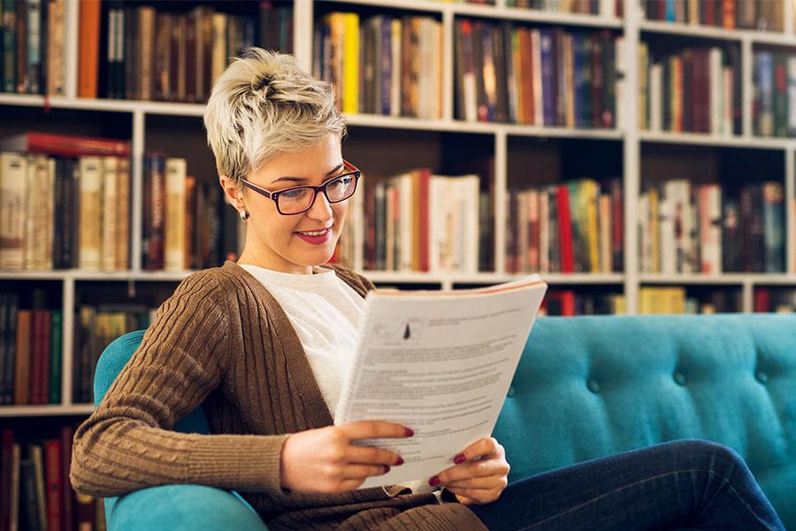 Photograph of a short-haired woman reading a printed document while seated on a teal sofa in a library.