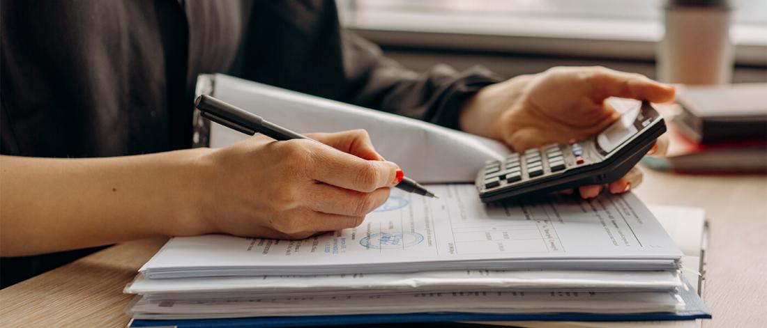 A close up shot of a woman's hands on top of a stack of documents with a calculator in hand.