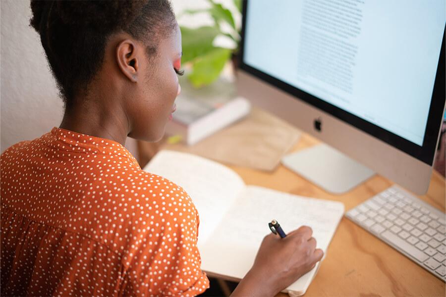 A woman writing on a notebook in front of a computer.