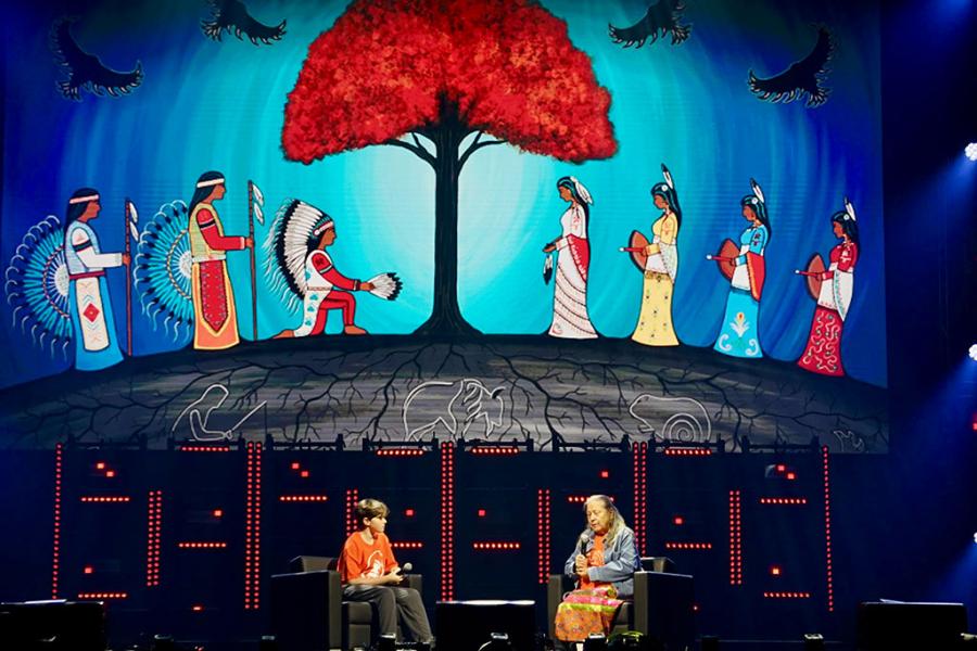 two people talking on stage under an indigenous illustration