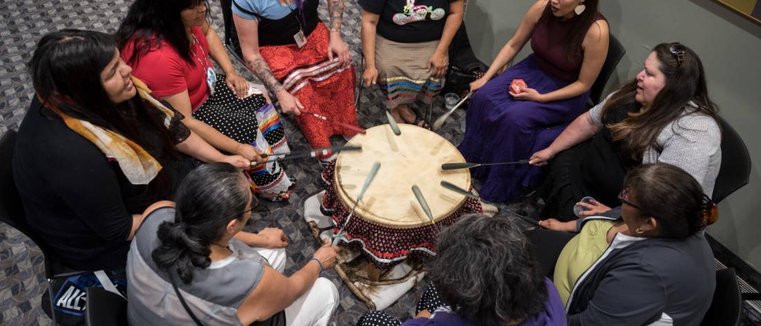 A group of women gathered in a circle beating a PowWow drum.