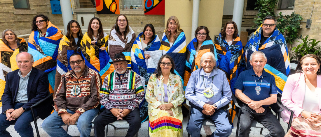 A group of people sit around a long table in a bright meeting room, smiling at the camera, with some making peace signs.