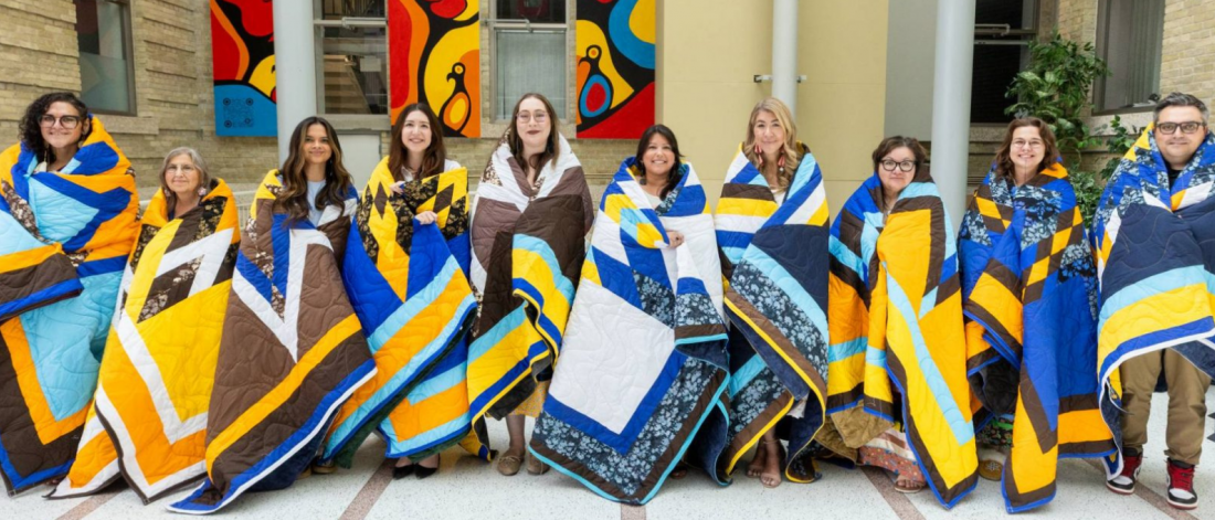 Ten people standing in a row, each wrapped in a colorful Indigenous Star Quilt, posing for a photo in a bright indoor hall.